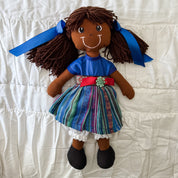 A handmade fabric doll with dark brown yarn hair and blue ribbon buns, wearing a blue top, a colorful patterned skirt, and black shoes, lying on a white quilted blanket.