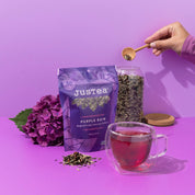 A styled product shot of Justea Purple Rain herbal tea. The scene features a purple pouch of the tea, a glass jar filled with loose herbal tea, a glass cup of brewed tea, a wooden spoon holding tea leaves, and a purple hydrangea flower on a solid purple background.