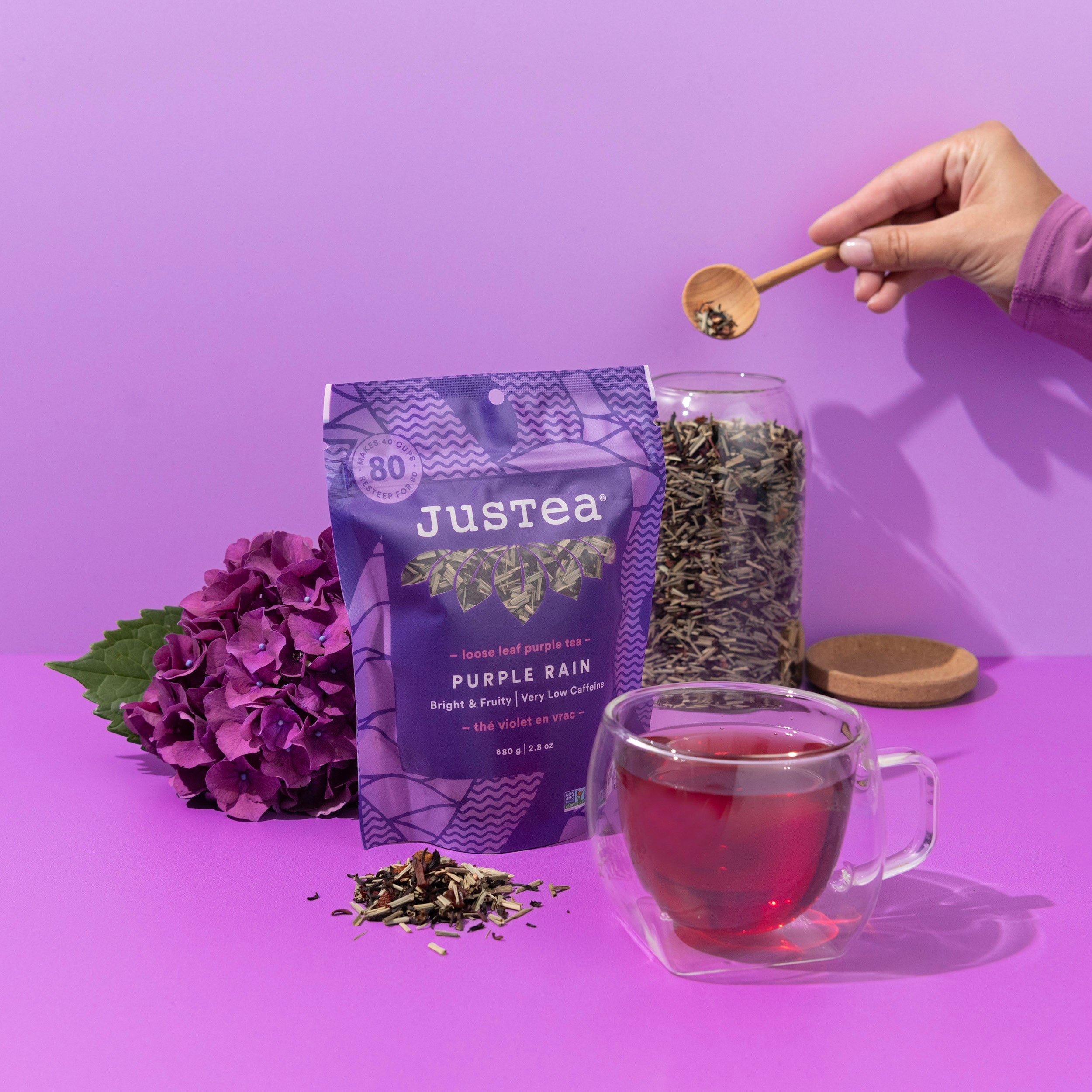 A styled product shot of Justea Purple Rain herbal tea. The scene features a purple pouch of the tea, a glass jar filled with loose herbal tea, a glass cup of brewed tea, a wooden spoon holding tea leaves, and a purple hydrangea flower on a solid purple background.