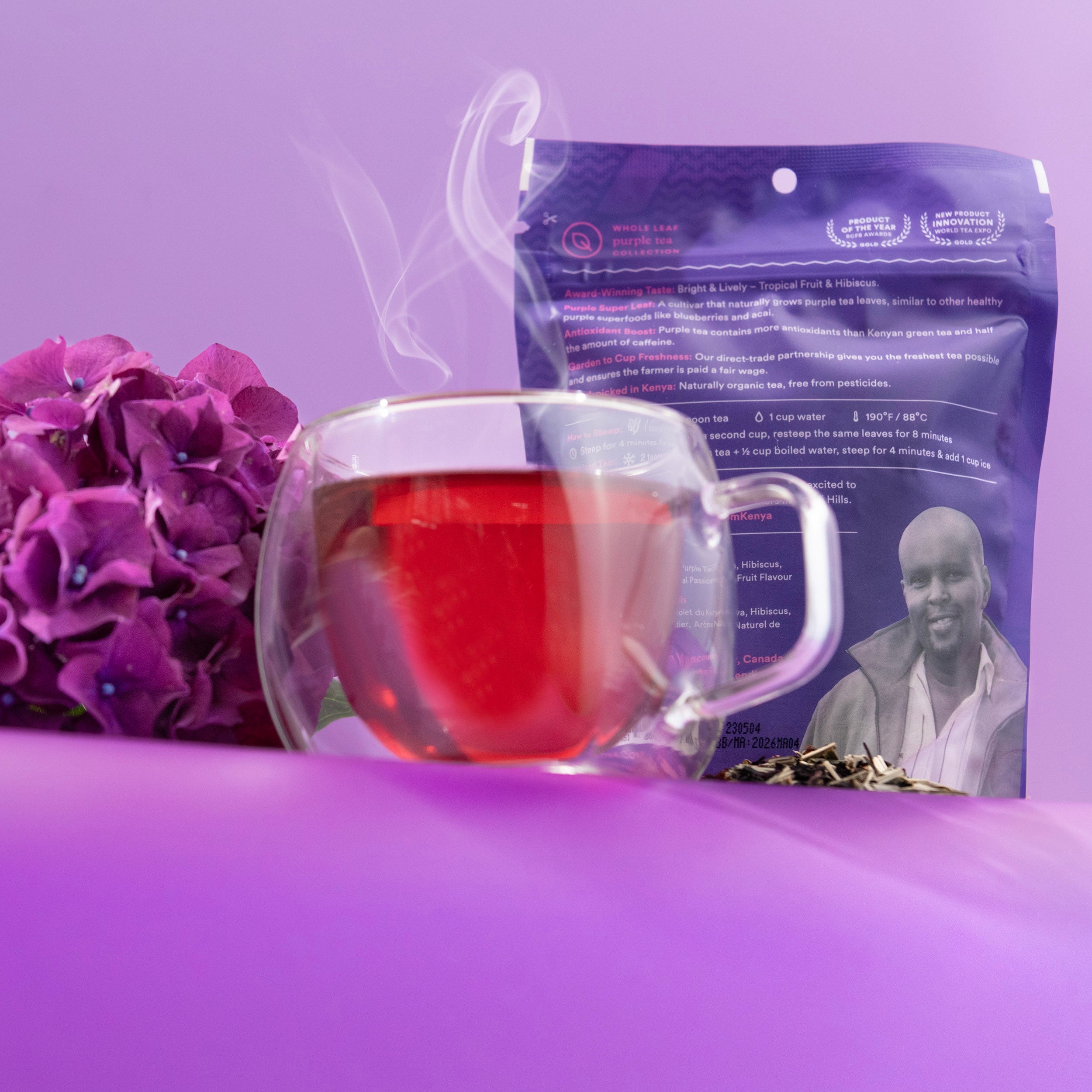 A glass mug filled with red tea sits in front of a purple stand-up pouch package, styled with purple hydrangea flowers and green leaves on a purple surface against a purple background.