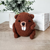 A brown crocheted beaver plush toy sitting on a wooden surface, with a white planter and green foliage behind it and a white wooden box to the right.