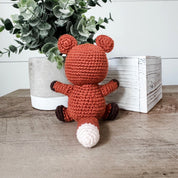 A crocheted plush toy fox in rust orange, brown, and white, sitting on a wooden table next to a white planter with green leaves and a white wooden box.