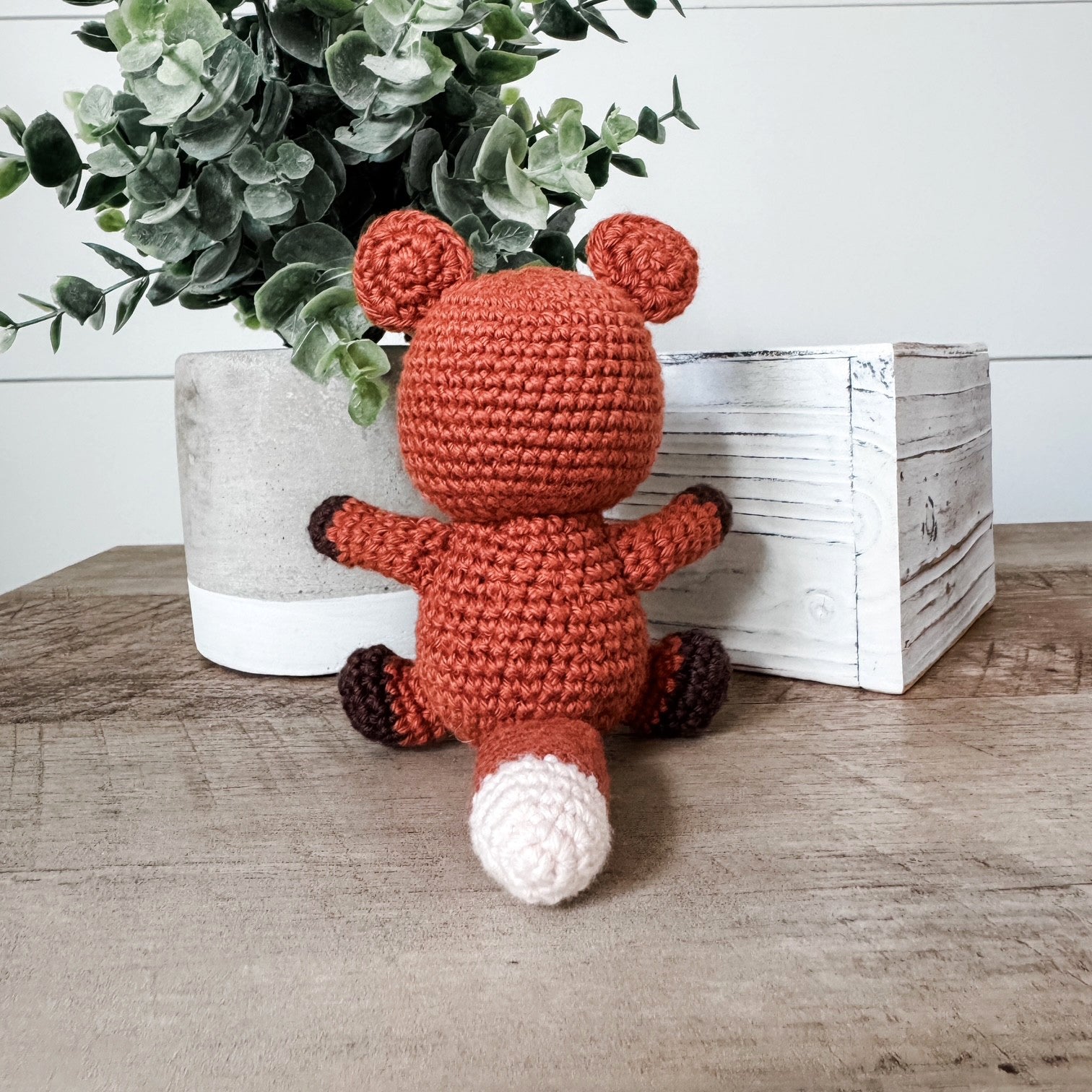 A crocheted plush toy fox in rust orange, brown, and white, sitting on a wooden table next to a white planter with green leaves and a white wooden box.