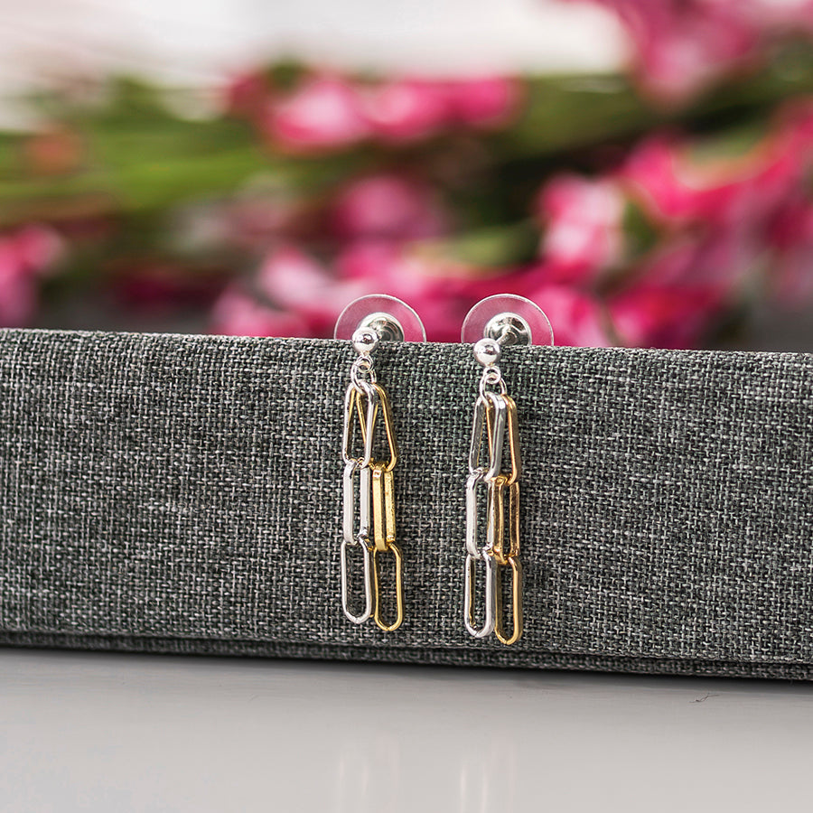 A pair of gold and silver paperclip drop earrings displayed on a textured gray jewelry stand, with a blurred pink floral background.