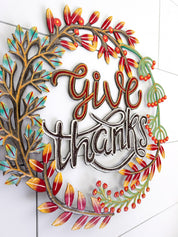 A decorative metal wall sign reading 'give thanks' in cursive script, surrounded by autumn-themed leafy branches in red, orange, yellow, teal, and green, mounted on a white tiled wall.