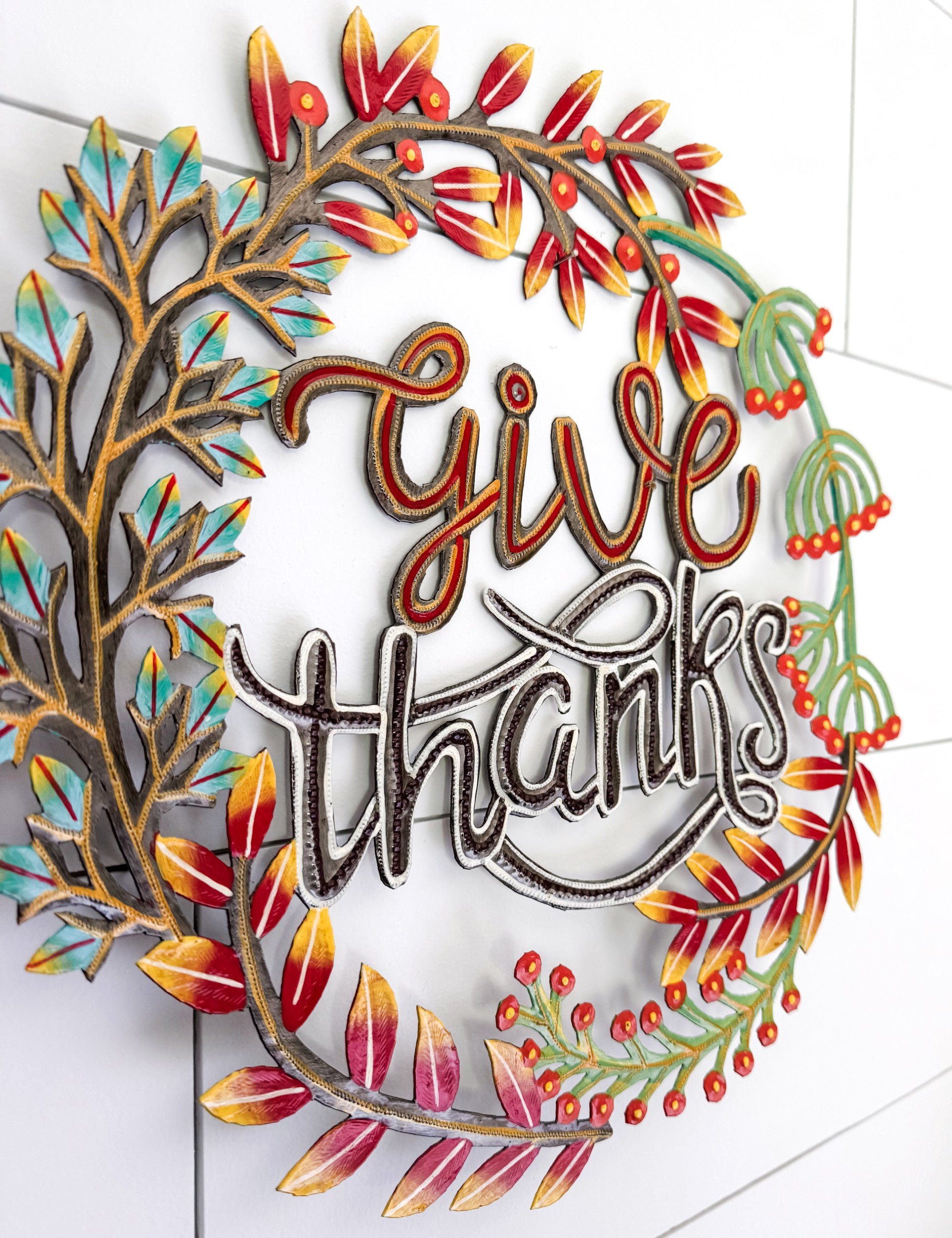 A decorative metal wall sign reading 'give thanks' in cursive script, surrounded by autumn-themed leafy branches in red, orange, yellow, teal, and green, mounted on a white tiled wall.