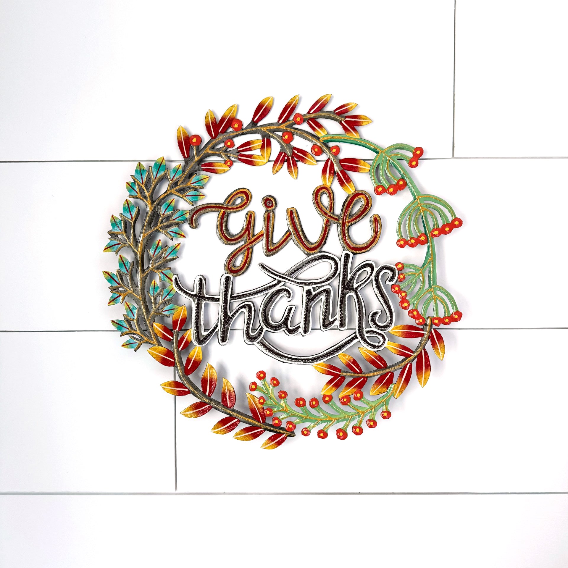 A circular metal wall sign with the words 'give thanks' in stylized lettering, surrounded by a wreath design with red berries and autumn leaves in red, orange, yellow, and green, mounted on a white paneled wall.