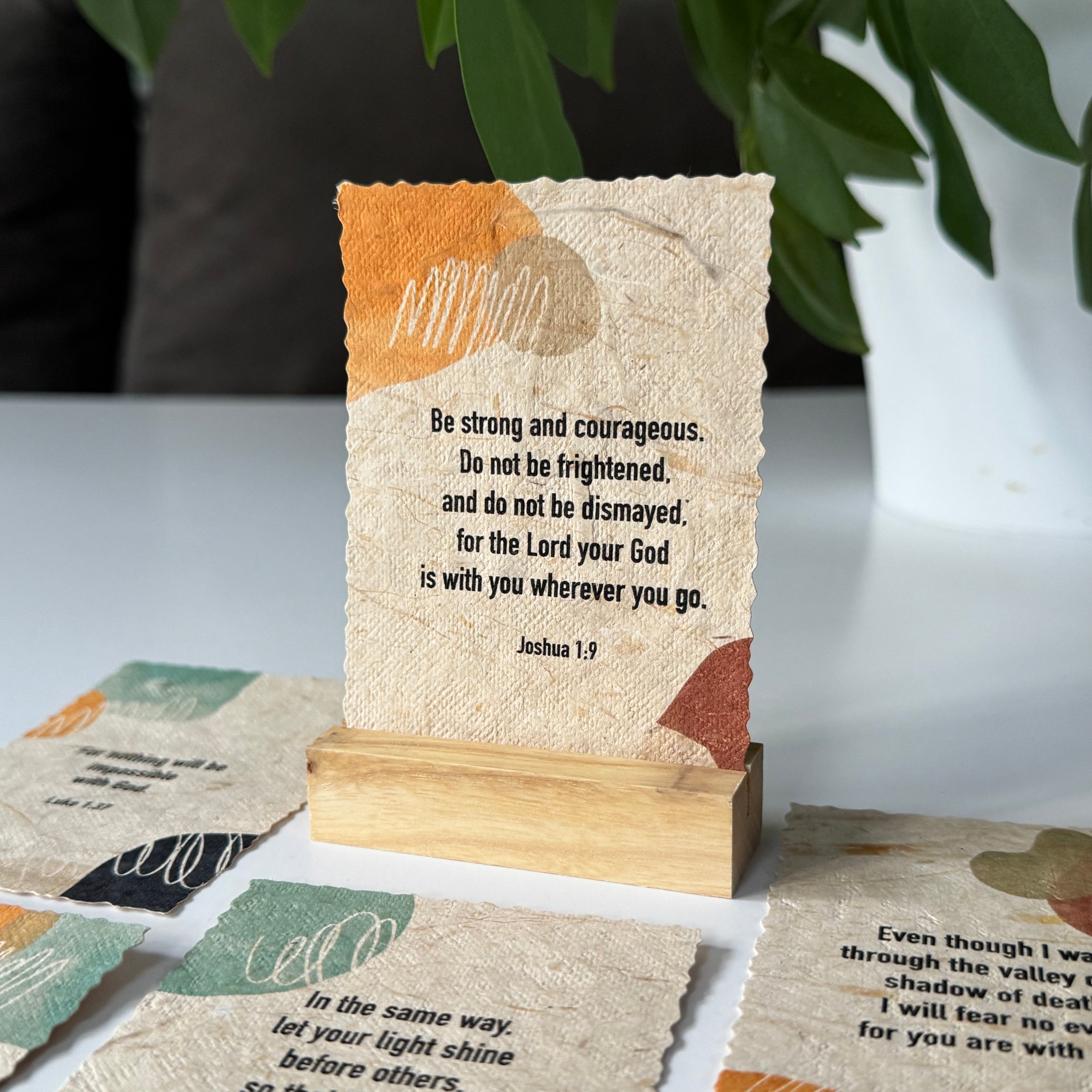 A set of textured, linen-like Bible verse cards displayed on a wooden stand, with additional cards spread out on a tabletop in a styled home setting with green leaves in the background.