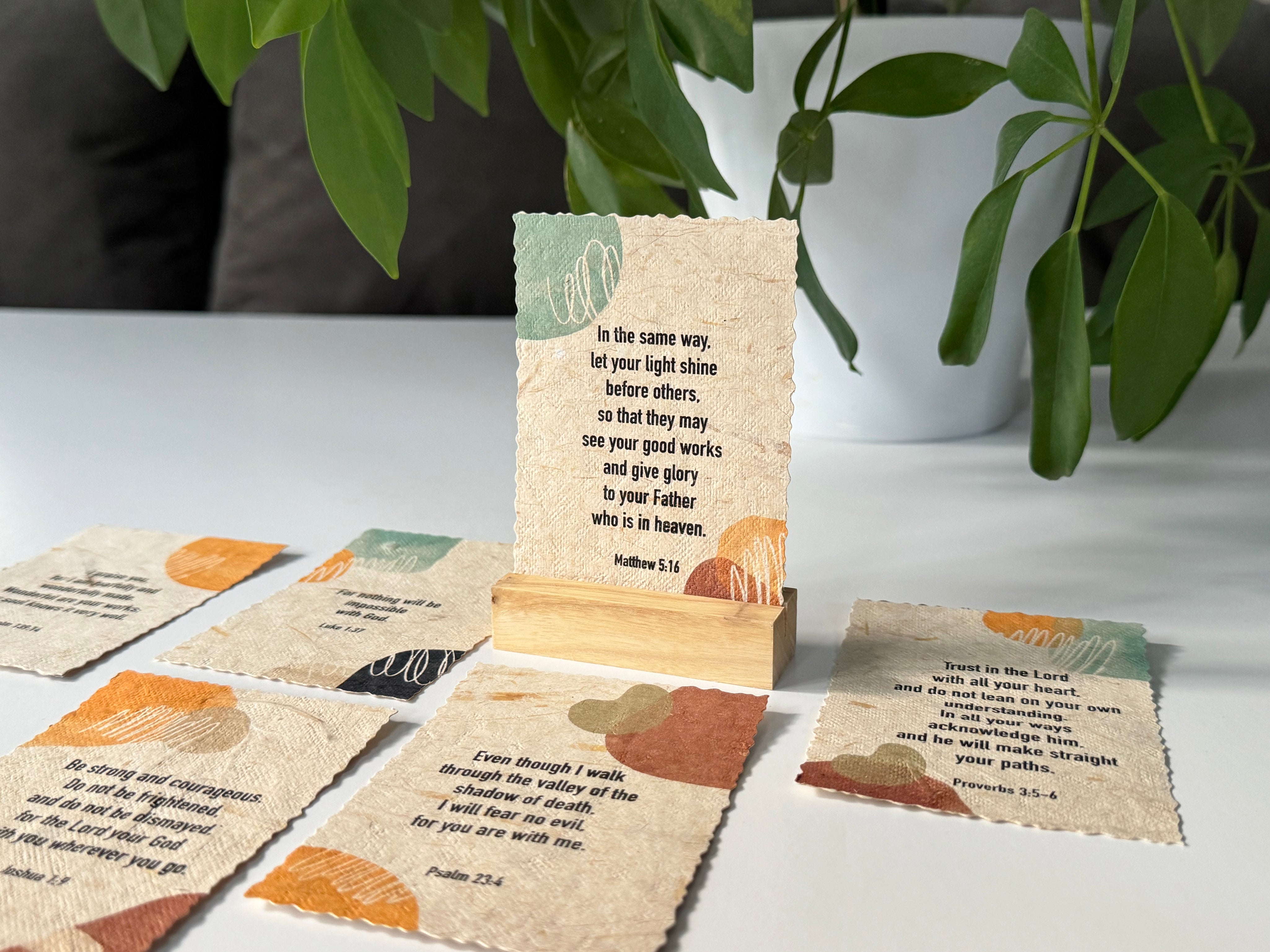 A set of small printed cards with scripture verses arranged on a tabletop, with one card standing upright on a wooden block. The cards have a textured beige background with orange and sage green abstract leaf-like shapes.