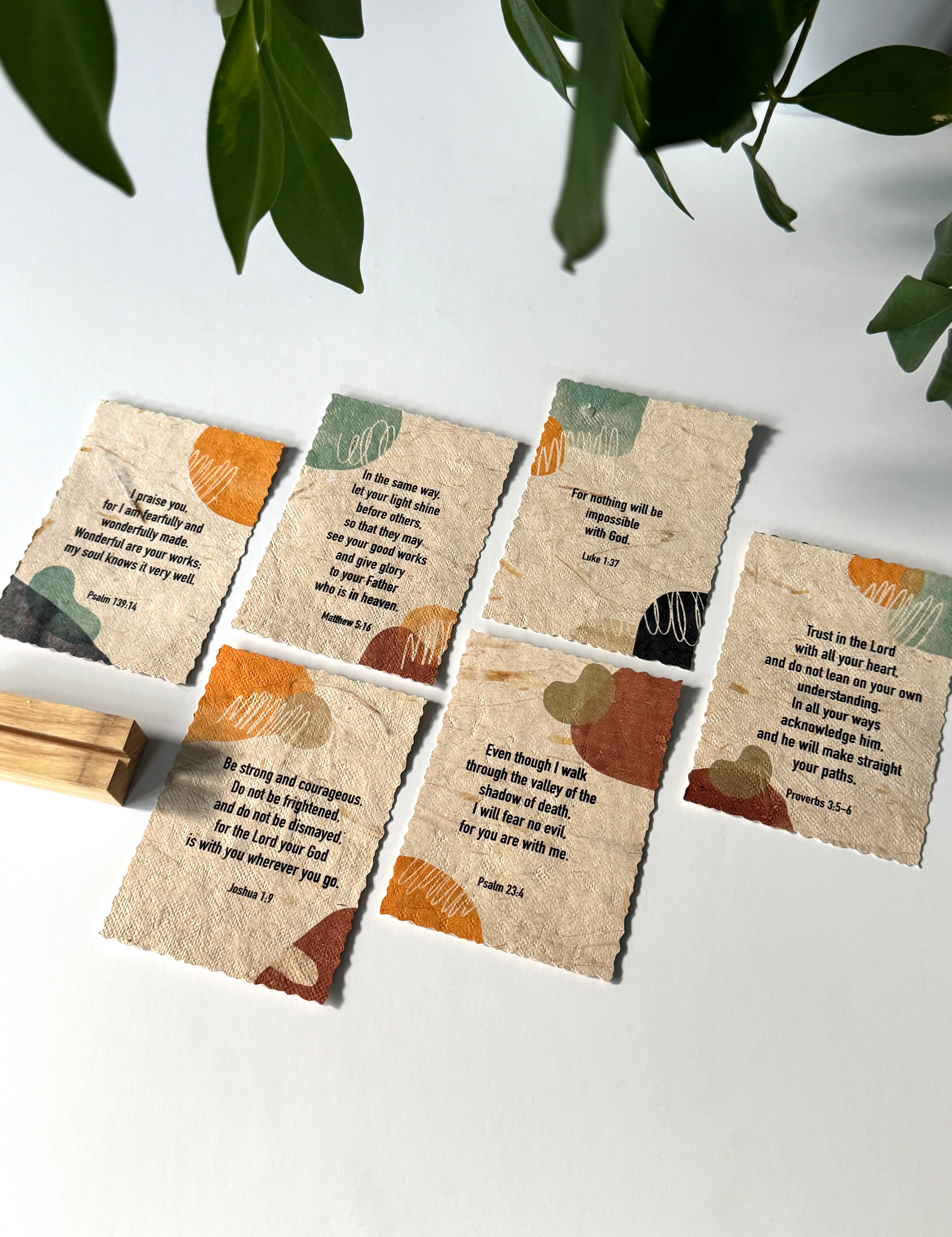 Six small square paper cards with torn edges, each printed with a Bible verse and abstract painted accents in orange, teal, brown, and black, arranged on a white surface with a wooden block and green leaves in the frame.