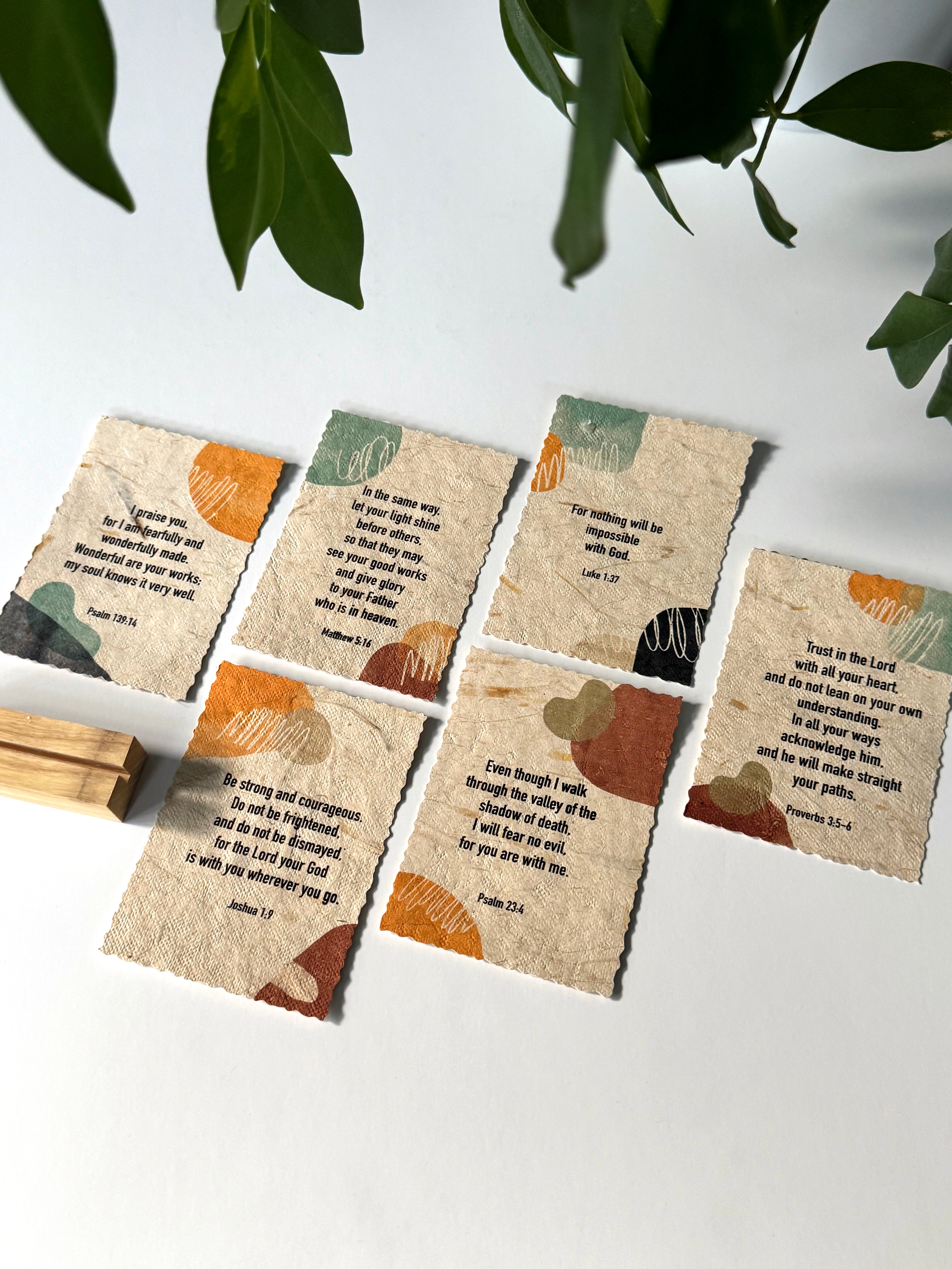 Six small square paper cards with torn edges, each printed with a Bible verse and abstract painted accents in orange, teal, brown, and black, arranged on a white surface with a wooden block and green leaves in the frame.