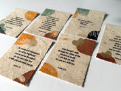 A set of small banana paper prayer cards with scalloped edges, printed with Bible verses and abstract earth-tone illustrations, arranged on a light surface.