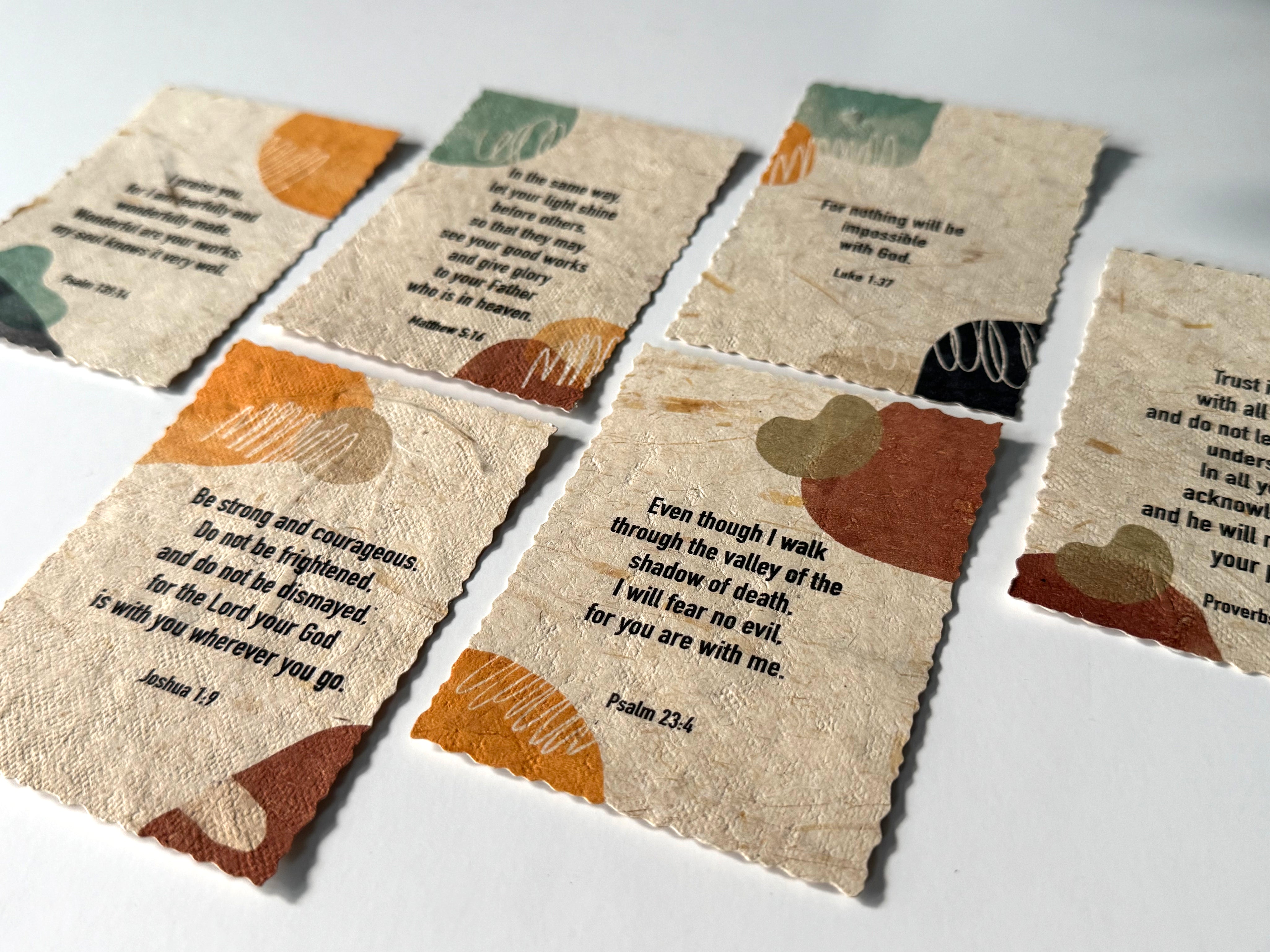 A set of small banana paper prayer cards with scalloped edges, printed with Bible verses and abstract earth-tone illustrations, arranged on a light surface.