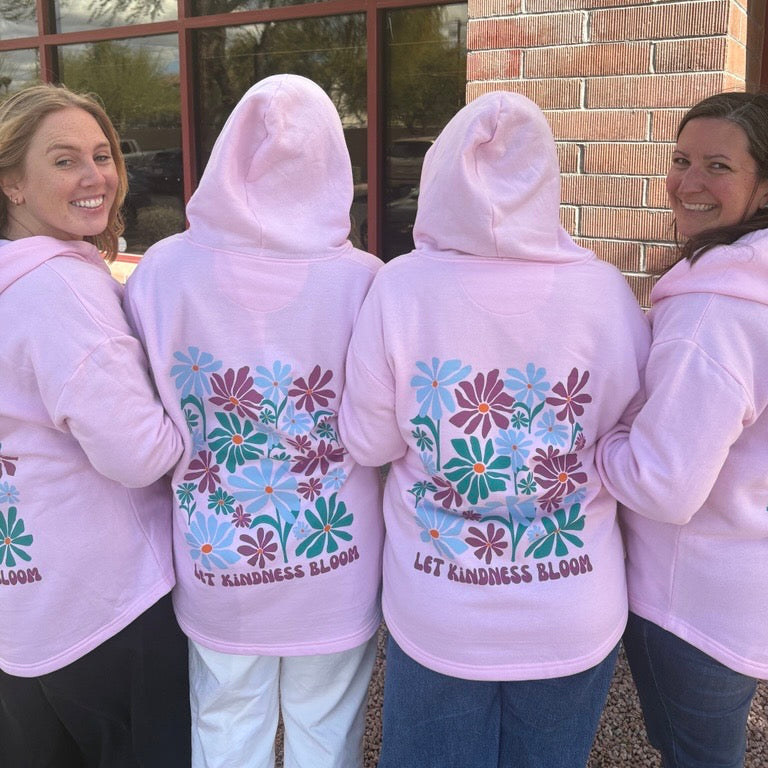 Four women wearing light pink hoodies with floral designs and 'Let Kindness Bloom' text.