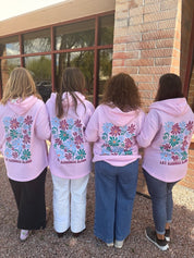 Four women wearing pink hoodies with floral designs and text on the back, standing outside a building.