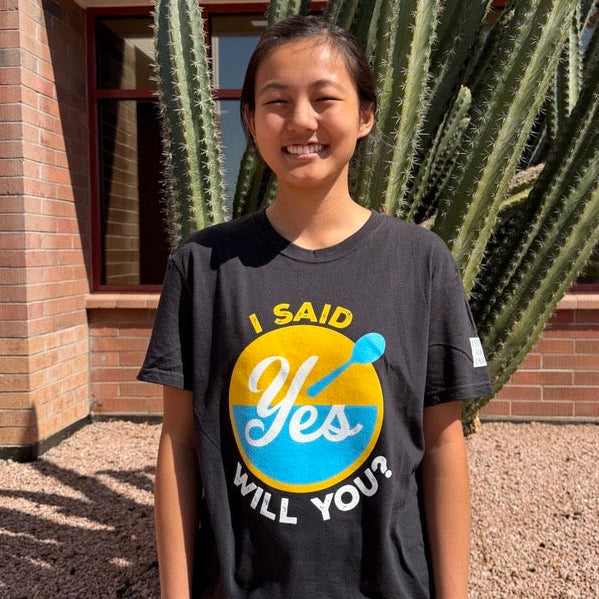 A smiling young woman stands outdoors in front of a brick building and tall cactus, wearing a black graphic T-shirt with a large yellow and blue circular design and the words 'I Said Yes Will You?'.