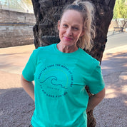Woman wearing a teal t-shirt with a graphic design, standing outdoors near a tree.