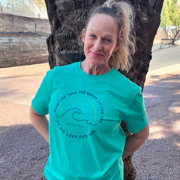 Woman wearing a teal t-shirt with a graphic design, standing outdoors near a tree.