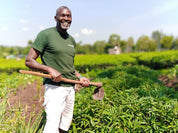 A smiling tea farmer wearing a green t-shirt and white pants stands in a lush green field holding a hand hoe.