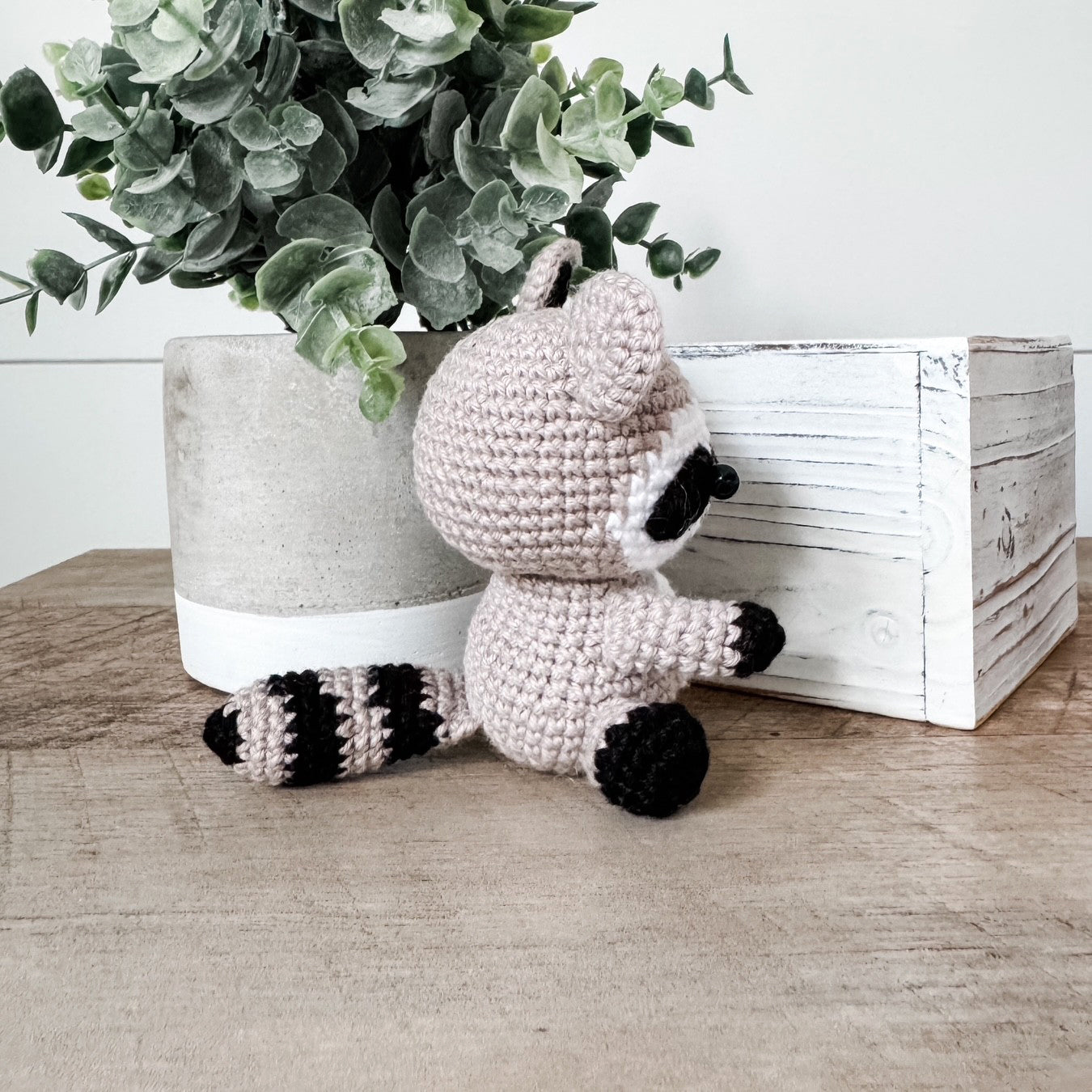 A crocheted raccoon toy sits on a wooden surface between a potted eucalyptus plant and a white wooden box. The raccoon is made of light blush pink yarn with black and white striped tail bands, black facial markings, and black paws.