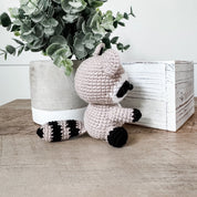 A crocheted raccoon toy sits on a wooden surface between a potted eucalyptus plant and a white wooden box. The raccoon is made of light blush pink yarn with black and white striped tail bands, black facial markings, and black paws.