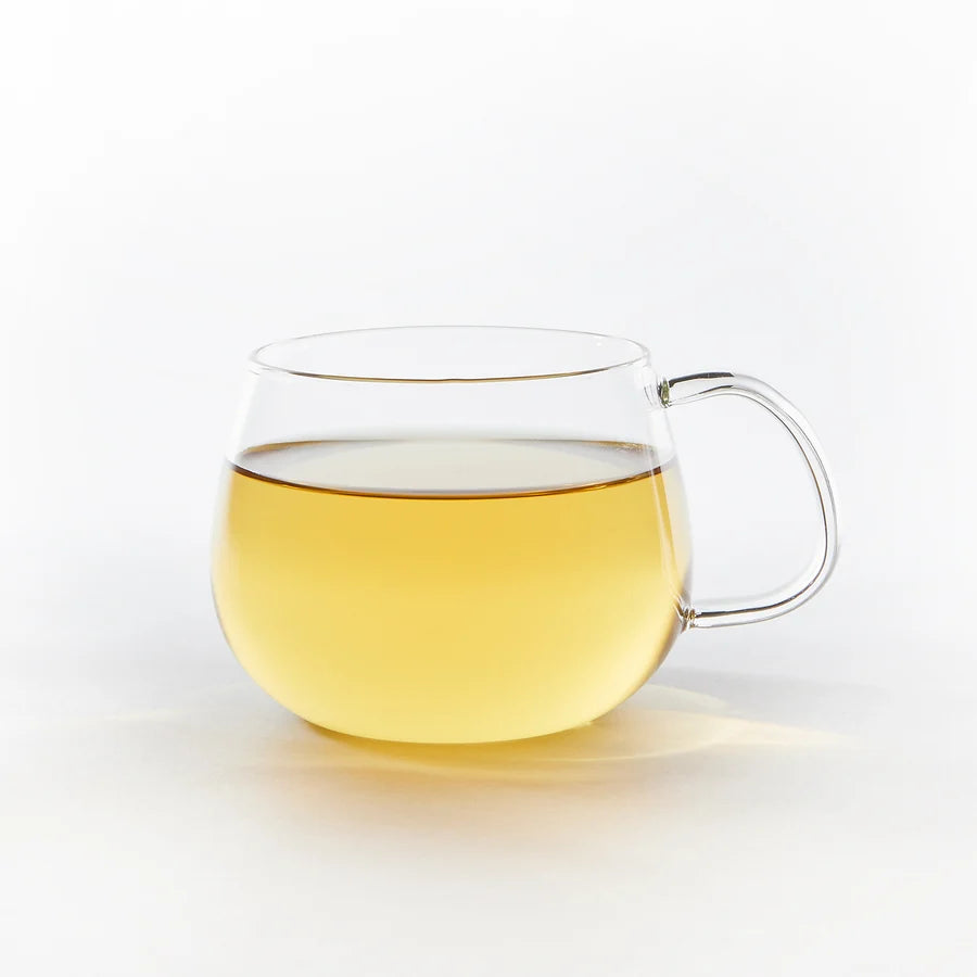 A clear glass teacup mug filled with pale golden tea, featuring a rounded bowl and a curved handle, set against a seamless white background.