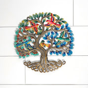 A colorful metal wall art piece shaped like a tree with roots, branches, and leafy canopy, decorated with multiple painted birds in red, orange, green, blue, and yellow. The piece is photographed against a white tiled wall.