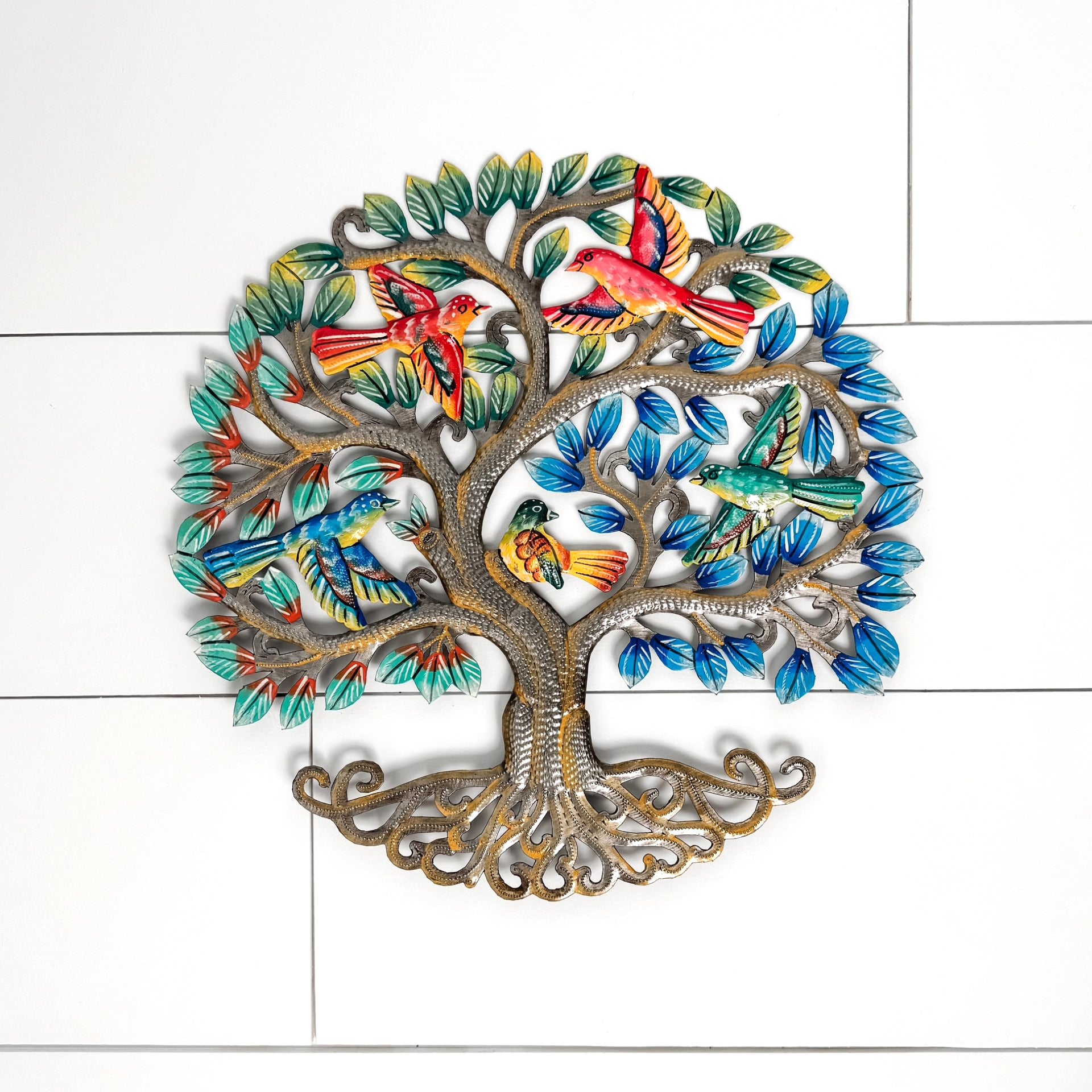 A colorful metal wall art piece shaped like a tree with roots, branches, and leafy canopy, decorated with multiple painted birds in red, orange, green, blue, and yellow. The piece is photographed against a white tiled wall.