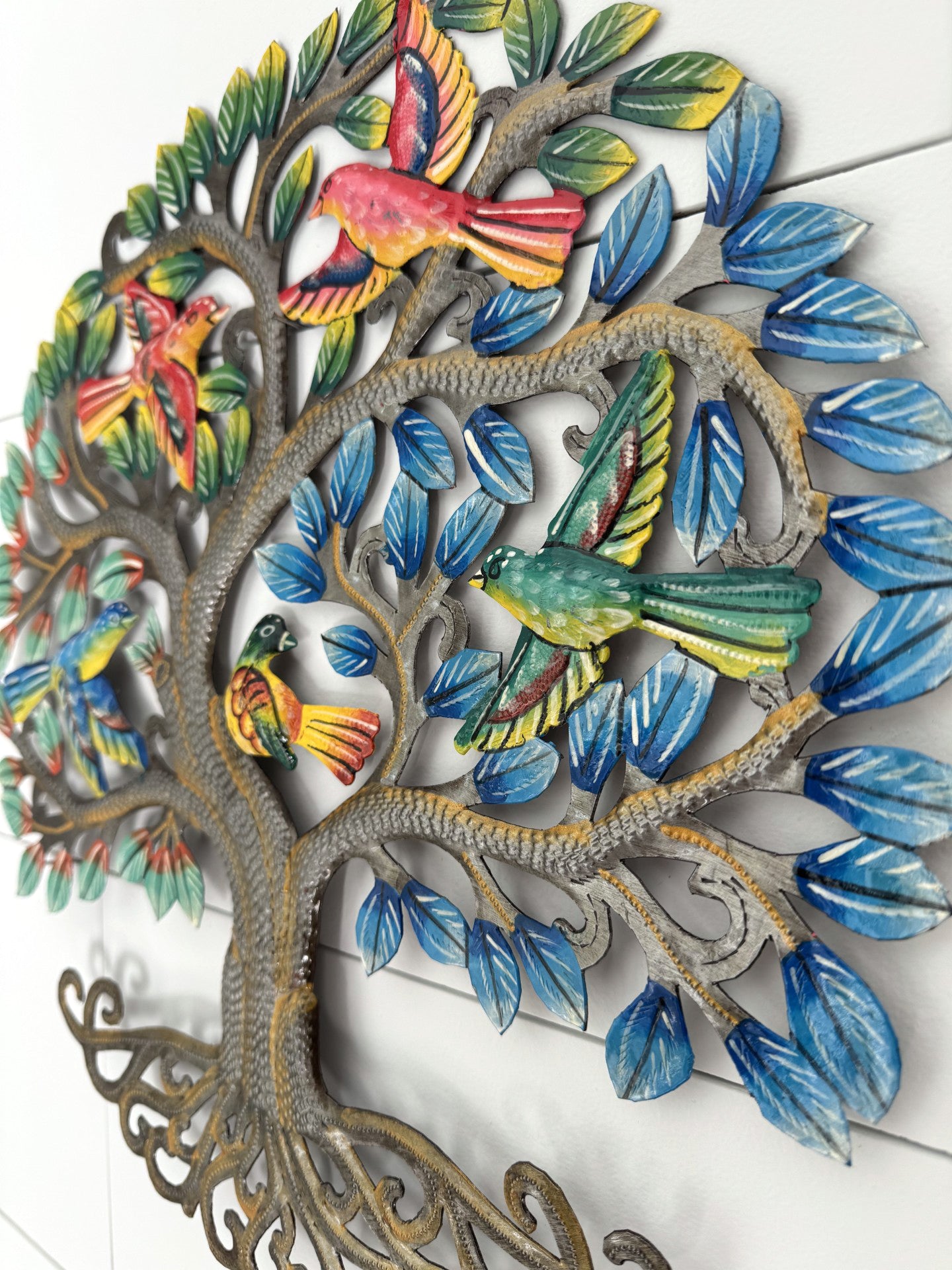 A decorative metal wall art piece shaped like a tree with colorful birds perched among blue leaves, shown against a white paneled wall.