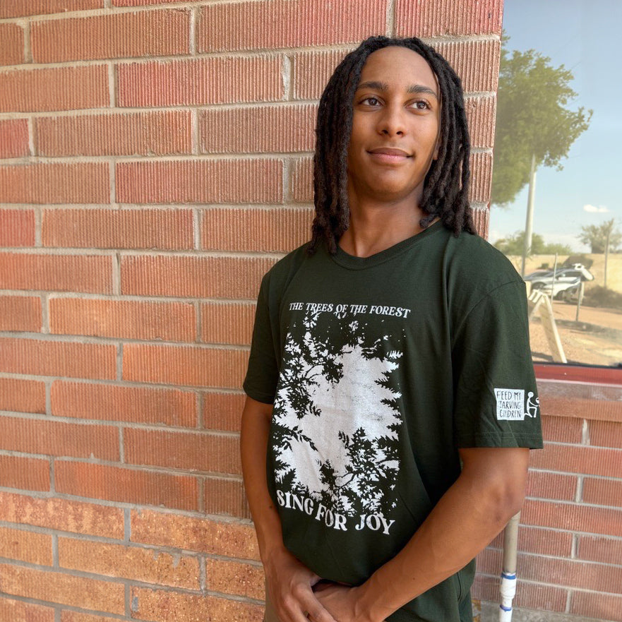 Person wearing a green t-shirt with a tree design and text, standing against a brick wall.