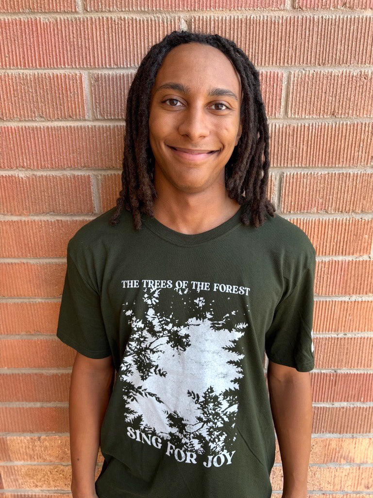 Person wearing a green t-shirt with a forest graphic and text, standing against a brick wall.