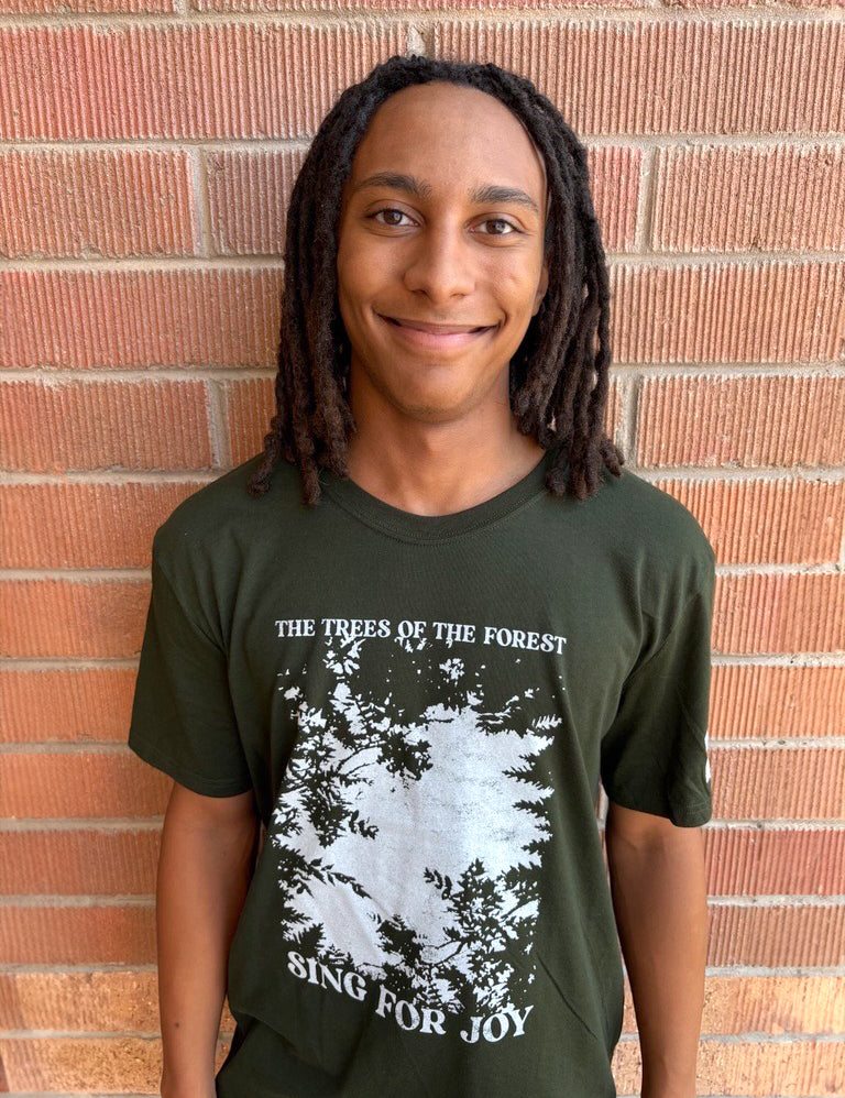 Person wearing a green t-shirt with a forest graphic and text, standing against a brick wall.