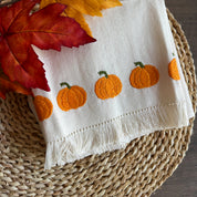 White towel with orange pumpkin embroidery on a woven mat with autumn leaves
