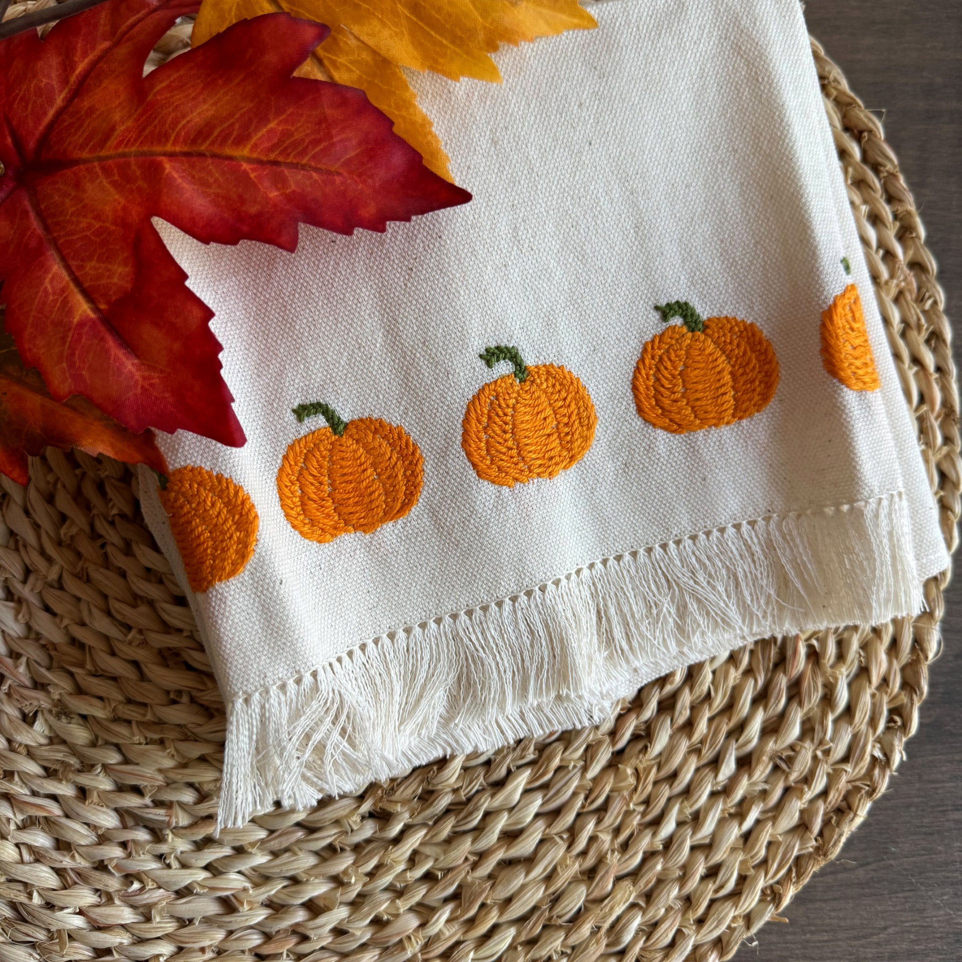 White towel with orange pumpkin embroidery on a woven mat with autumn leaves